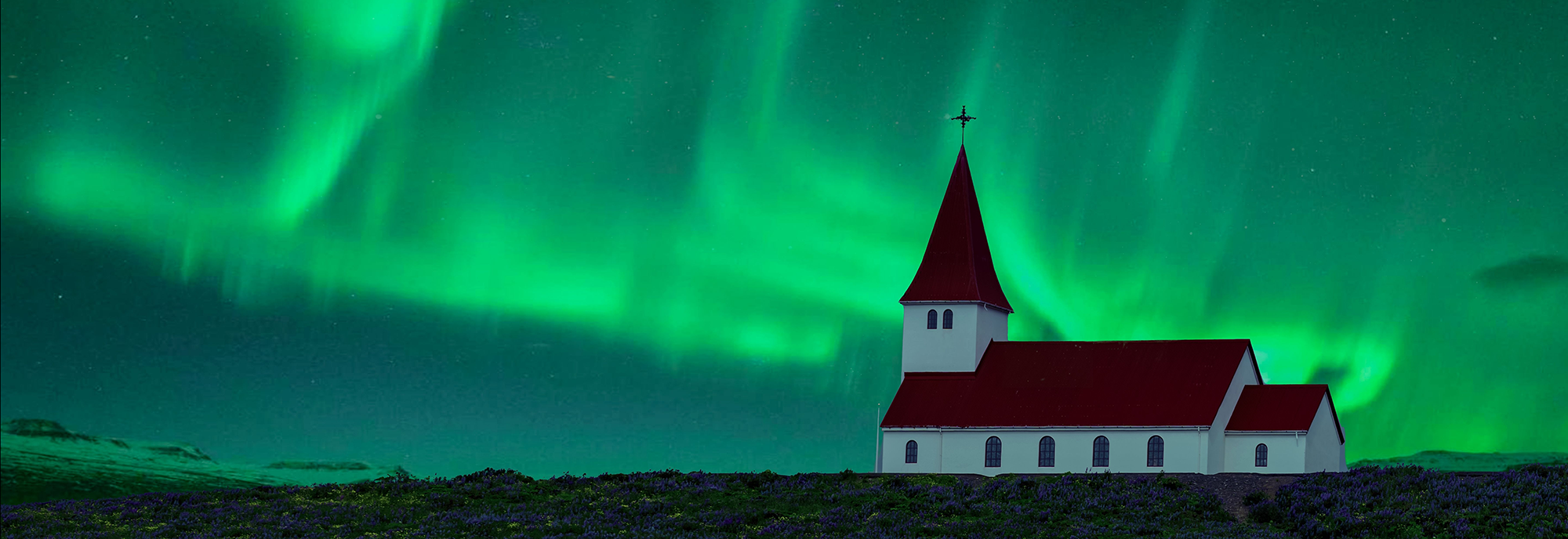 Een wit houten kerkje onder een felgroene lucht vol golvend Noorderlicht in een donker IJslands landschap.